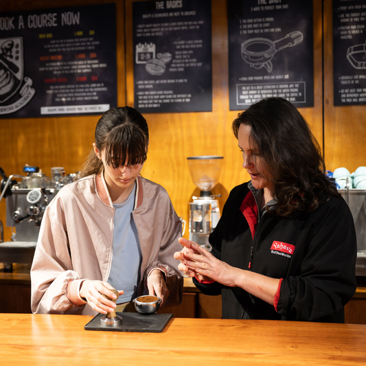 A woman in a black jacket with a red logo and white text stands at a wooden counter, holding a small cup while a younger woman in a beige jacket pours coffee. The background shows a warm wood-paneled wall with black posters and coffee equipment.